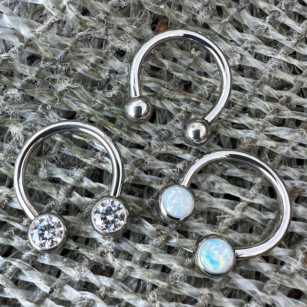 Three threadless titanium circular barbells, with cubic zirconi faceted gem ends, white opal cabochon gem ends, and titanium ball ends.